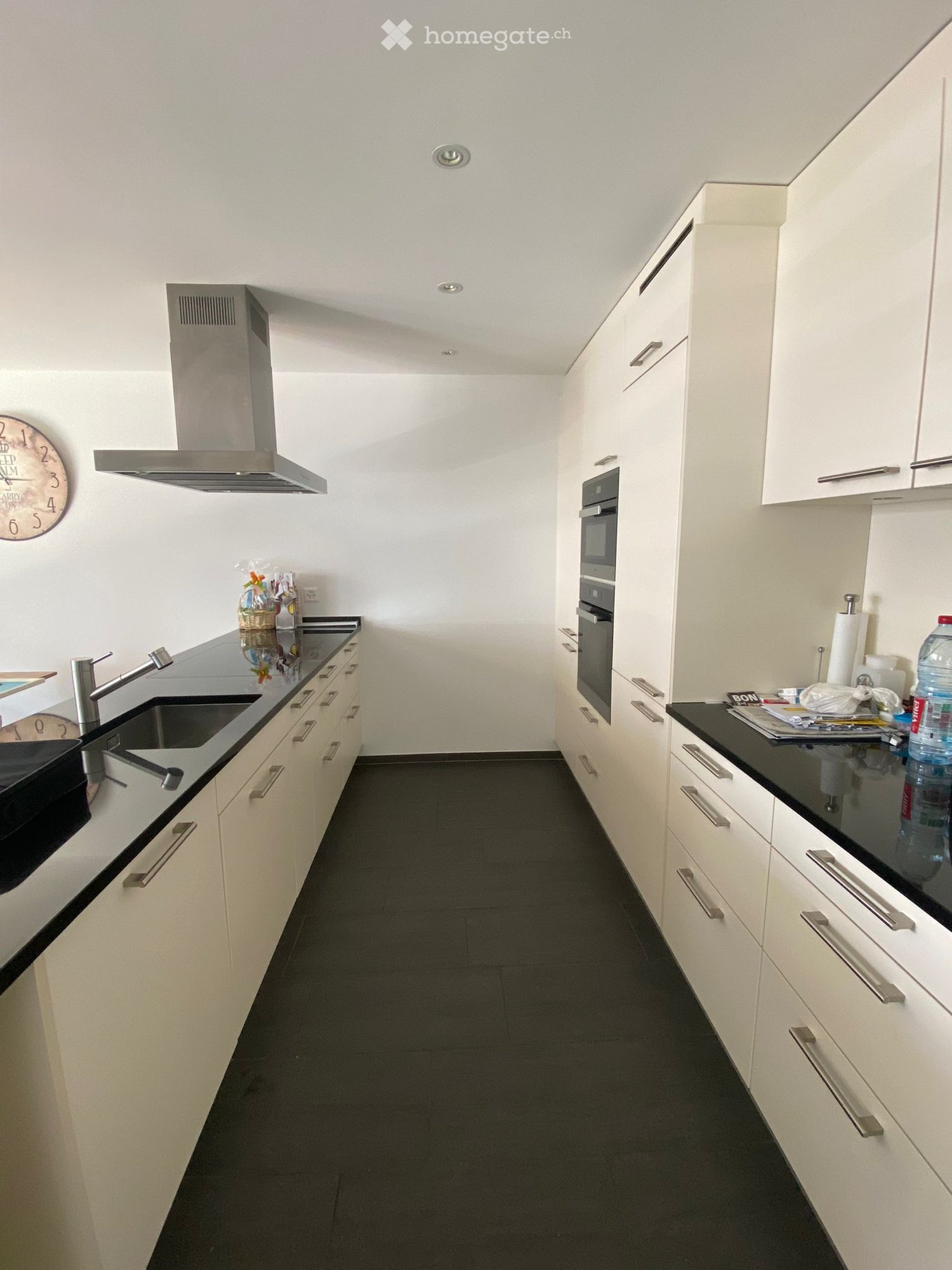 Modern kitchen with white cabinets, black countertops, stainless steel sink, and built-in oven. Features a sleek design with a dark tiled floor.