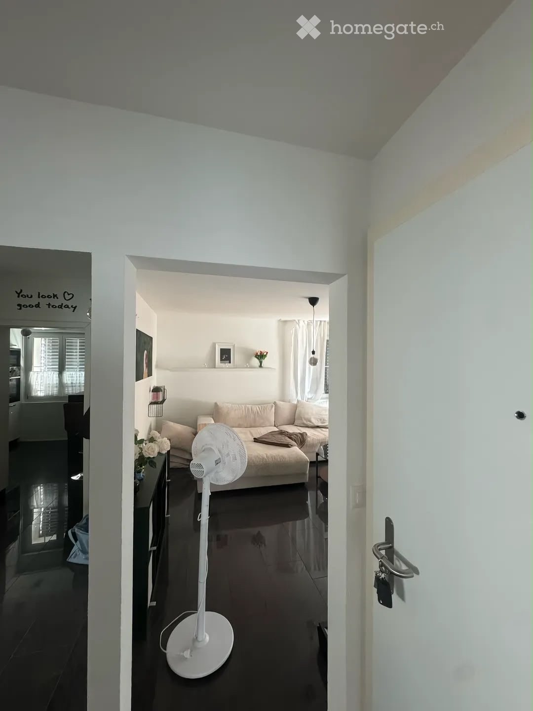 A modern living room with a beige sofa, a white standing fan, a small table with a flower vase, and a wall-mounted picture frame. The room features white walls, dark flooring, and a large window with white curtains.