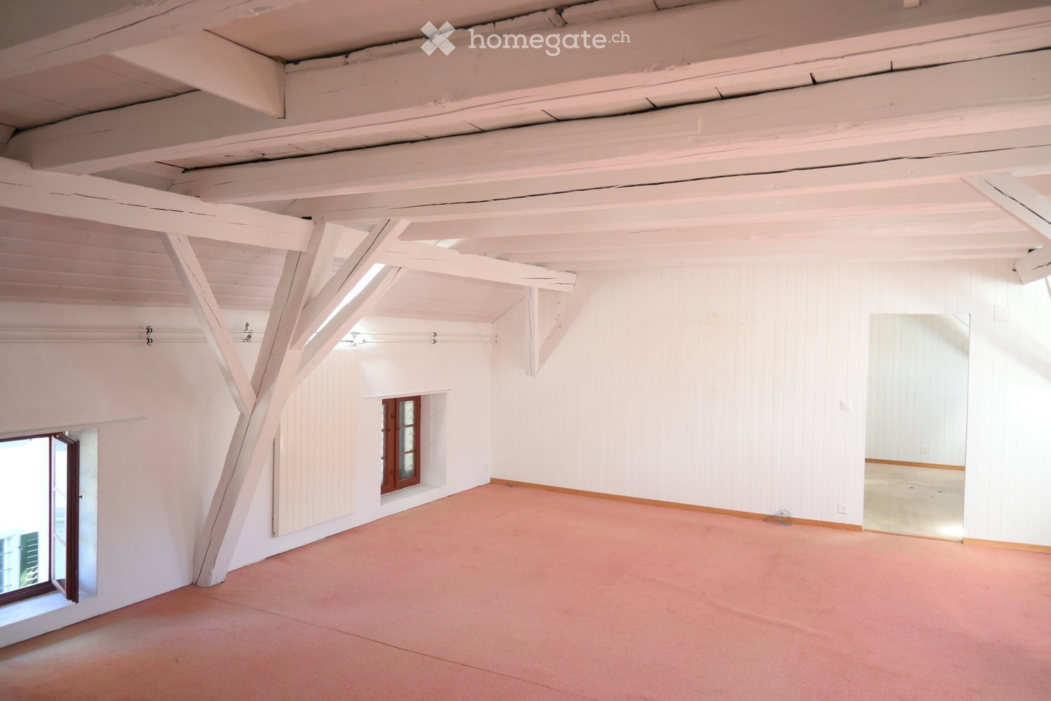Empty room with white walls, exposed wooden beams, and a pink carpeted floor. Features include a large window on one side and a smaller windowed door leading to another space.