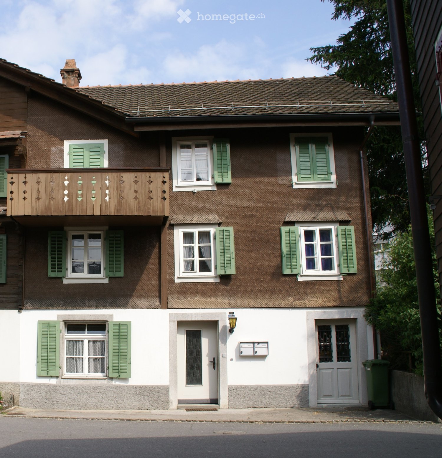 Two-story house with brown tiled roof, green shutters, white walls, and a wooden balcony on the upper floor.