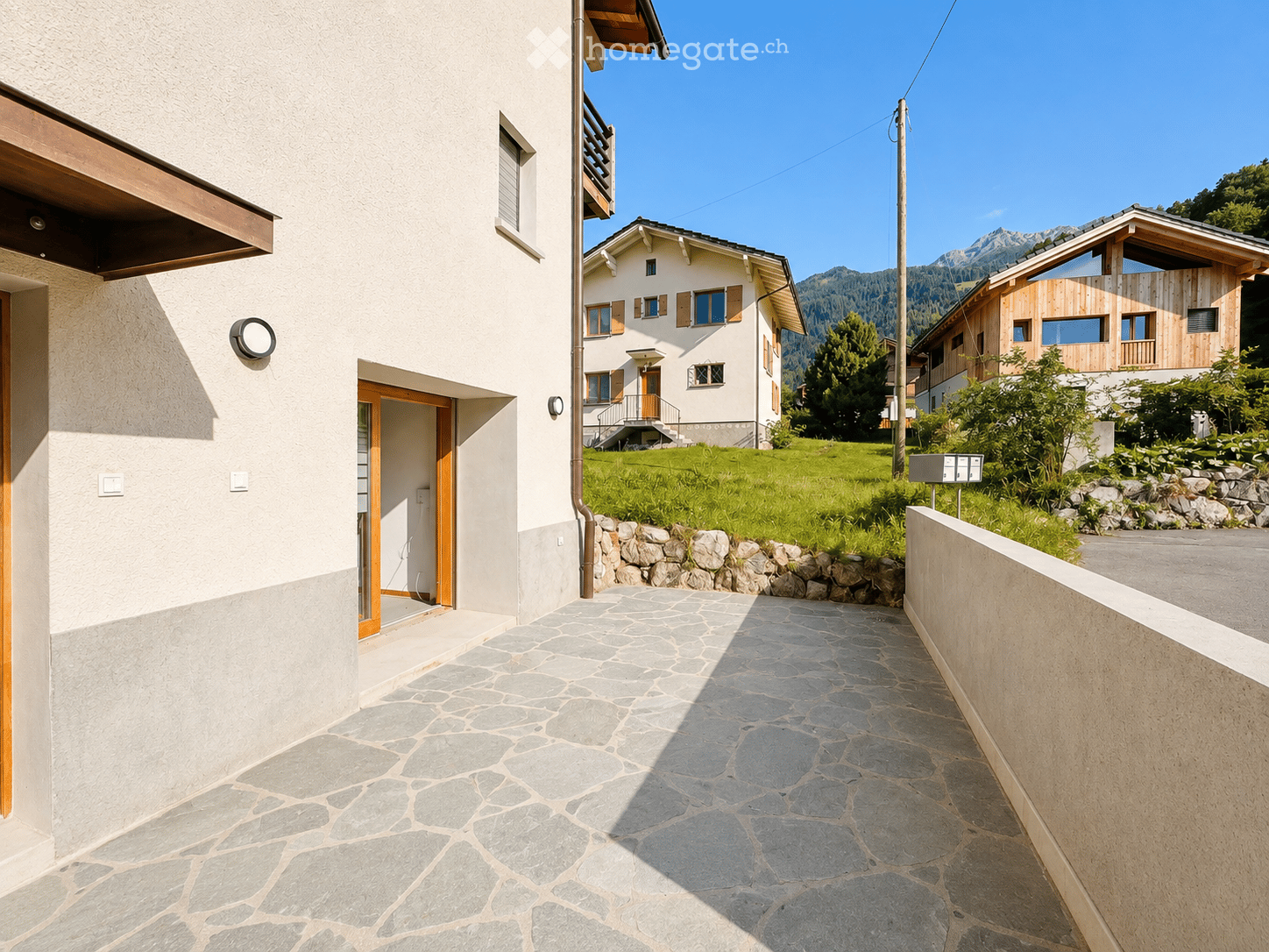 A stone-paved pathway leads to a modern house with a white facade and wooden accents, surrounded by greenery and mountains in the background.
