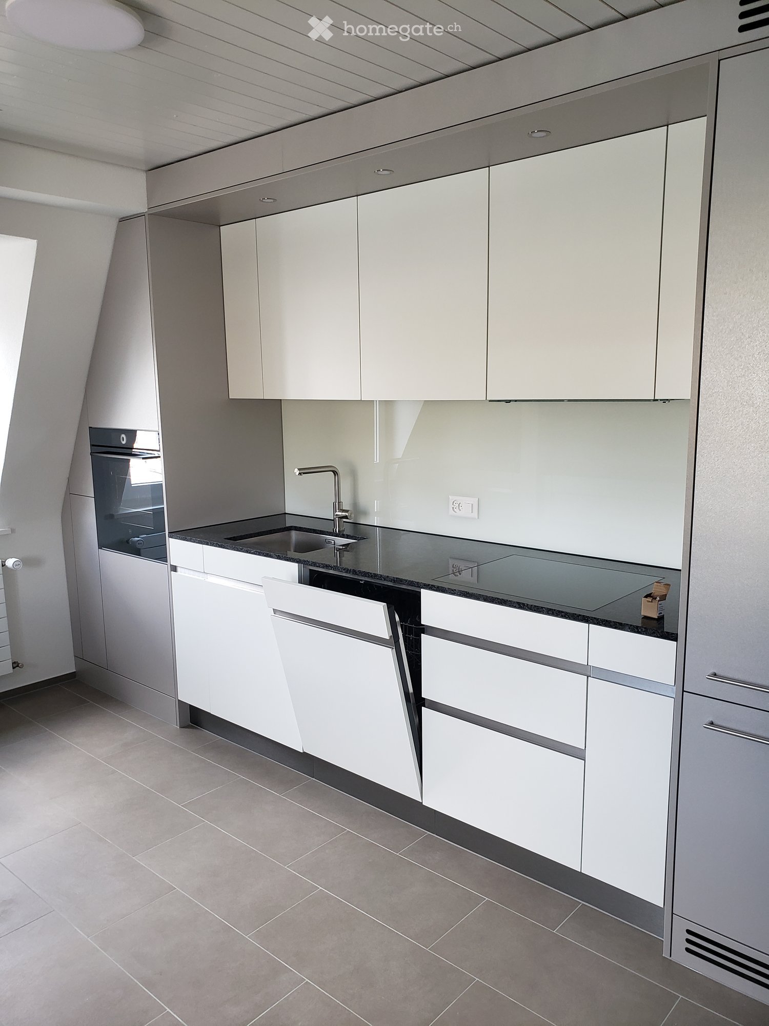 Modern kitchen with white cabinets, stainless steel sink, and built-in oven. Features a sleek, minimalist design with a dark countertop and tiled floor.