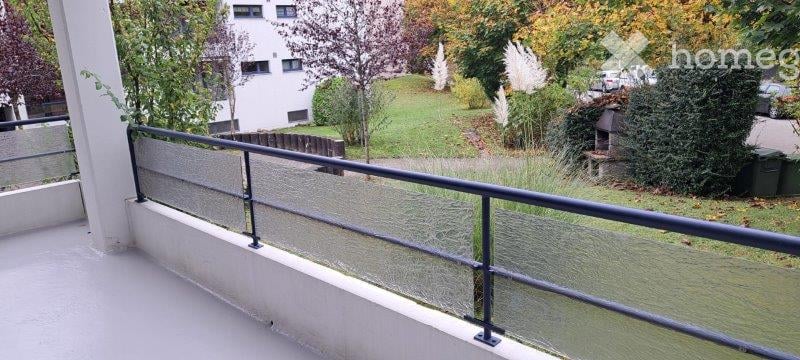 Balcony with glass railing, overlooking a green lawn and trees, with a building in the background.