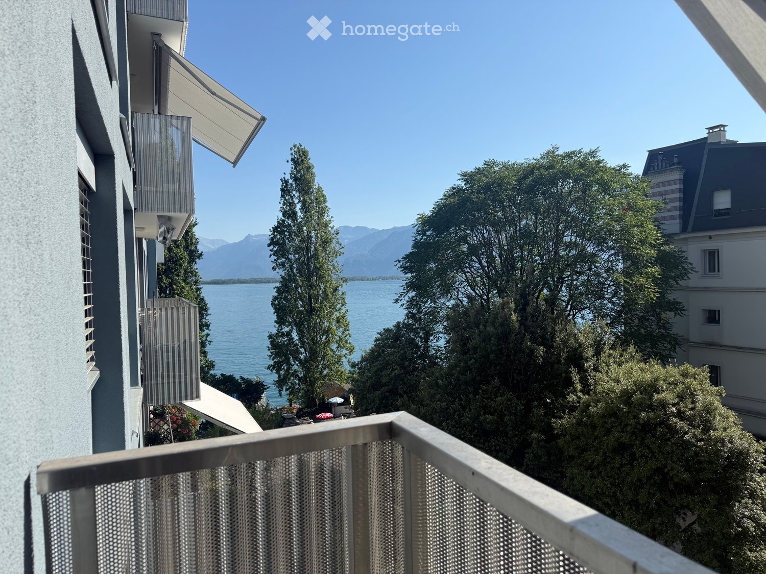 Balcony overlooking a scenic lake with mountains in the background, surrounded by trees and a neighboring building.