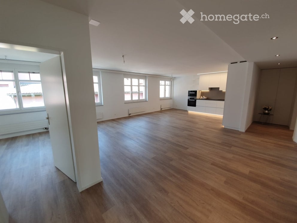 Bright, empty room with wooden flooring, white walls, large windows, and a small kitchen area visible in the background.