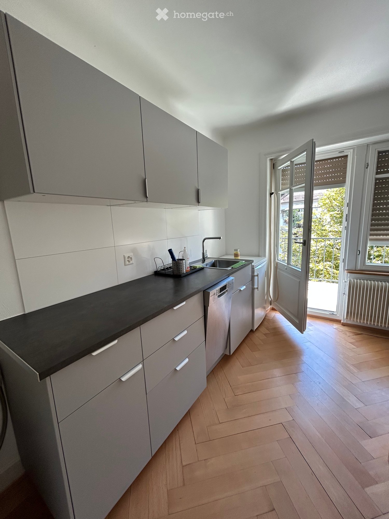 Modern kitchen with light grey cabinets, black countertop, wooden flooring, and a dishwasher. An open door leads to a balcony with greenery visible outside.