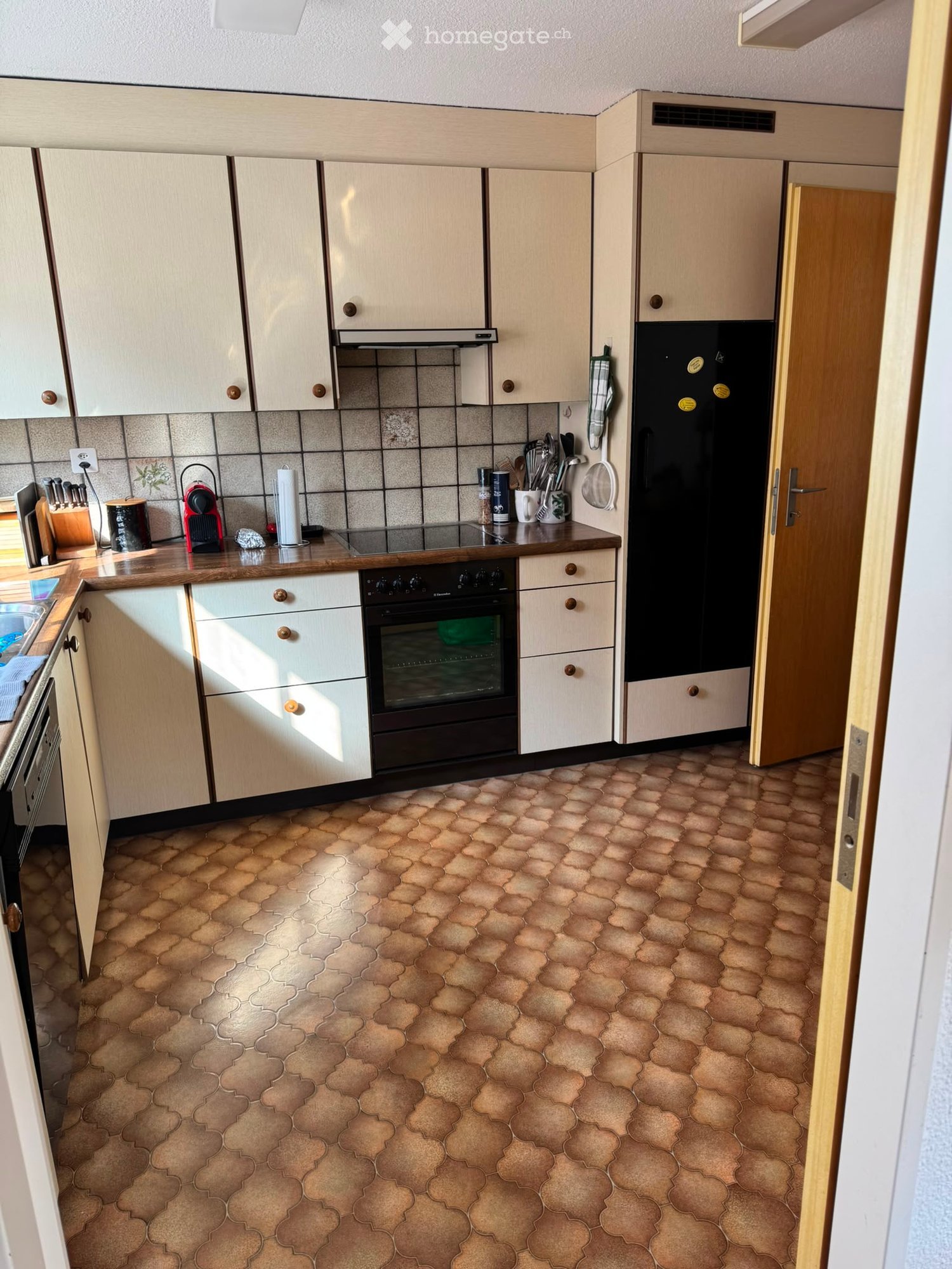 A well-equipped kitchen with light wood cabinets, a black refrigerator, a built-in oven, and various appliances on the counter, including a coffee maker and a knife block. The floor features a patterned tile design.