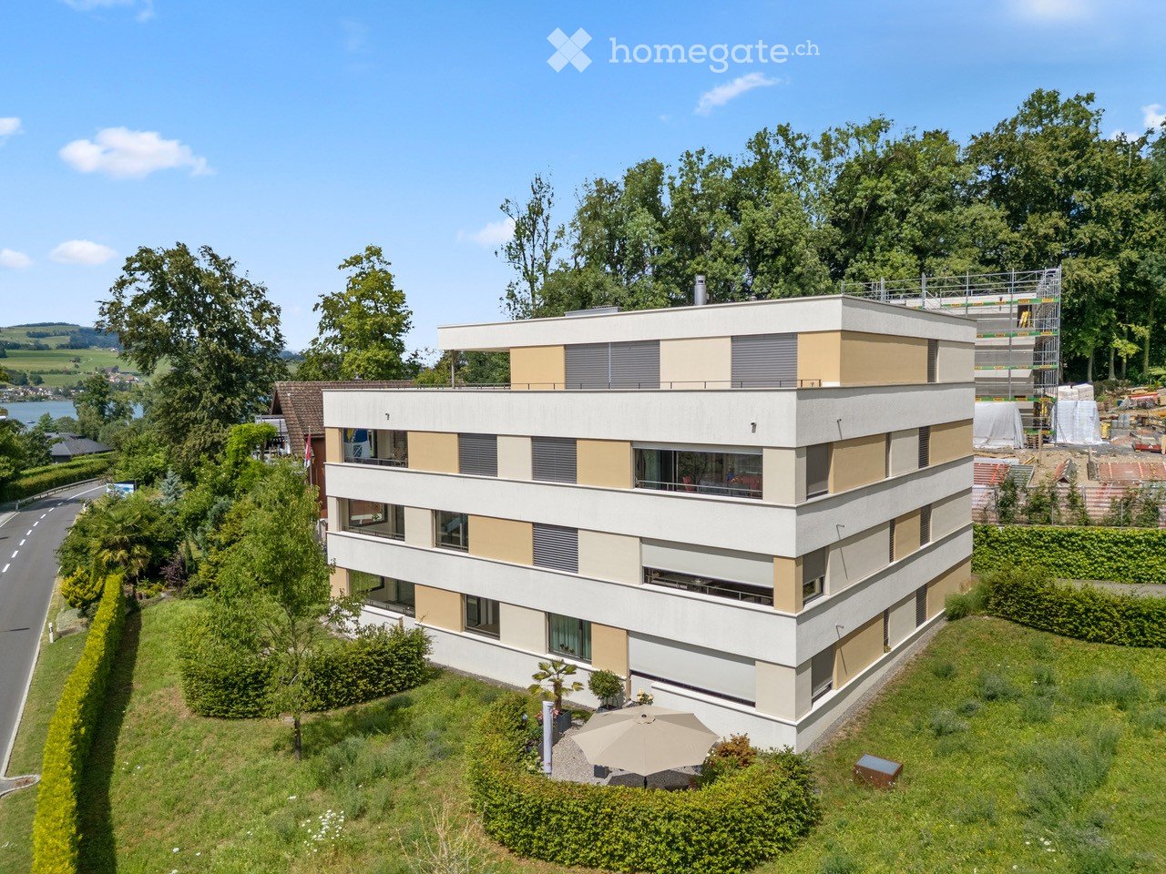 Modern multi-story building with white and beige facade, large windows, and balconies. The surrounding area includes a well-maintained lawn, trees, and a nearby road.