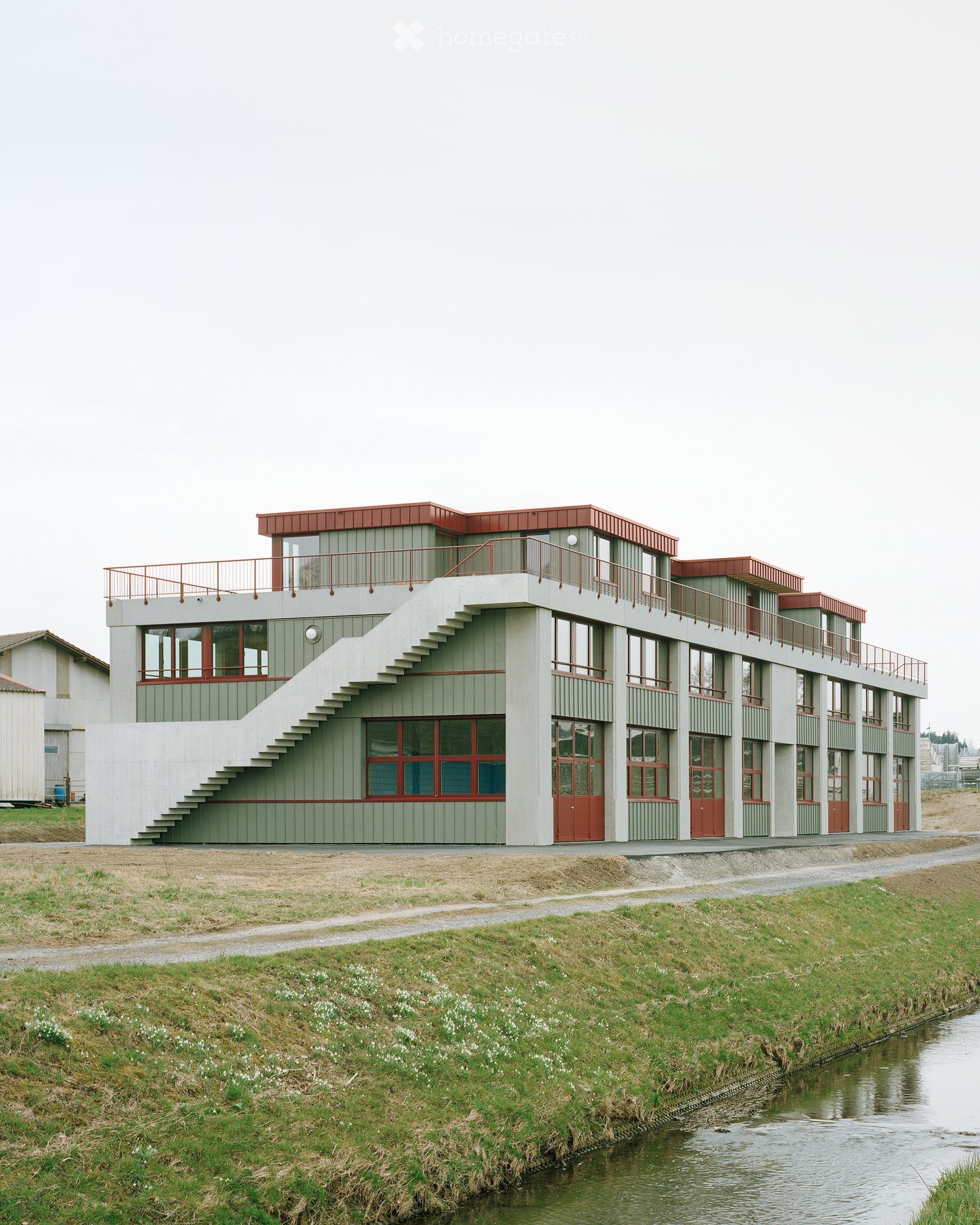 Modern two-story building with green siding, red trim, and a red roof, featuring a staircase leading to an upper-level balcony.
