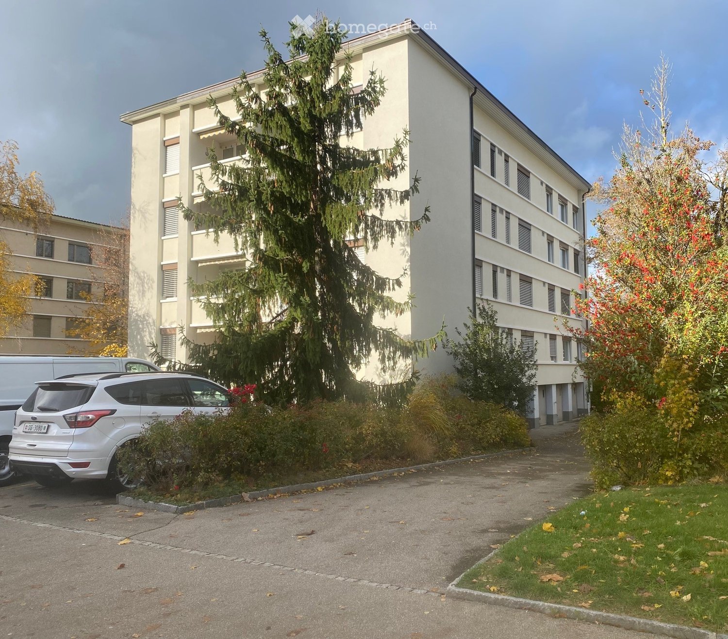 Multi-story residential building with balconies, surrounded by trees and parked cars.