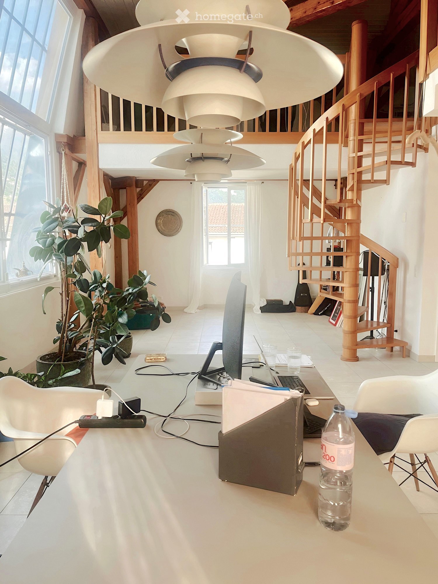 Bright, modern living space with a large white pendant light fixture, a staircase leading to an upper level, and a workspace area with a laptop, screen, and keyboard on a table. There are white chairs, a plant, and a water bottle on a small table.