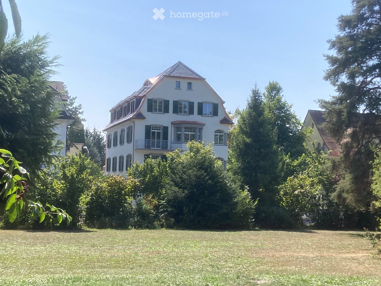 A two-story white house with a red-tiled roof, multiple windows, and a small balcony, surrounded by lush greenery and trees.