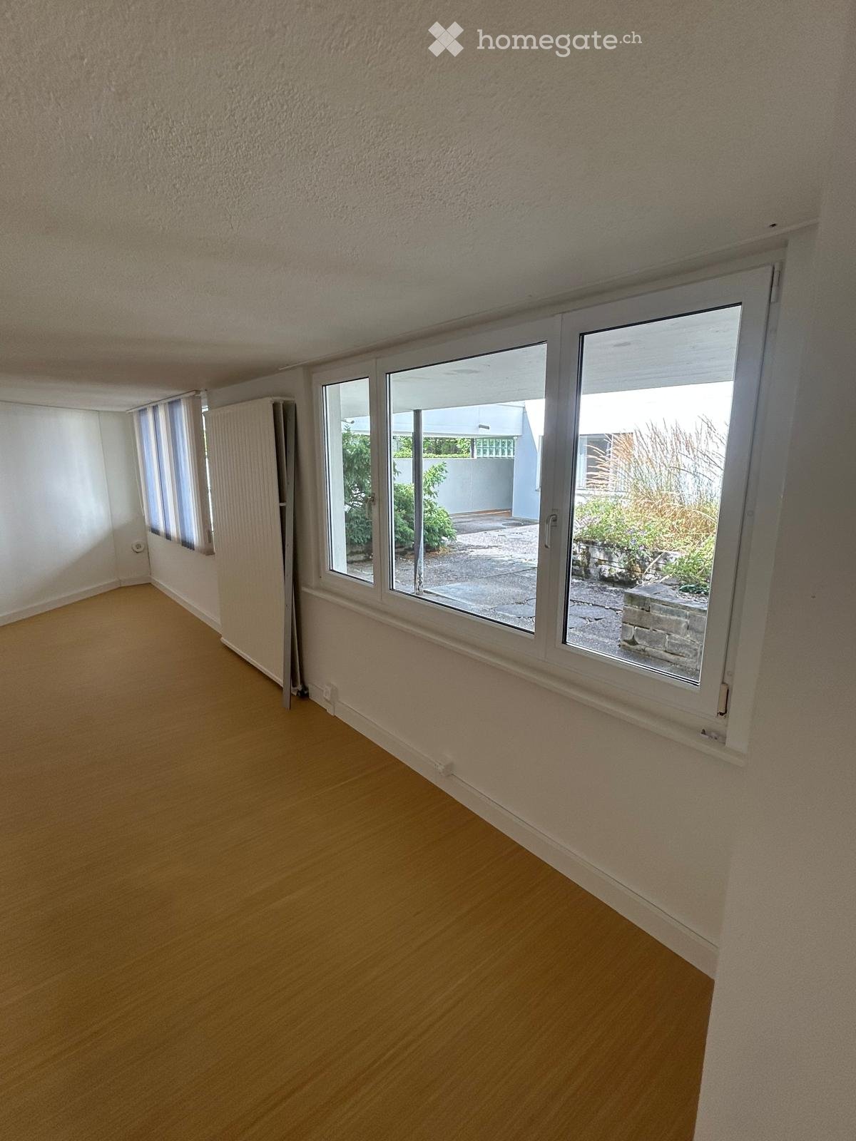 Empty room with wooden flooring, white walls, and large windows offering a view of an outdoor patio area with plants and a paved surface.