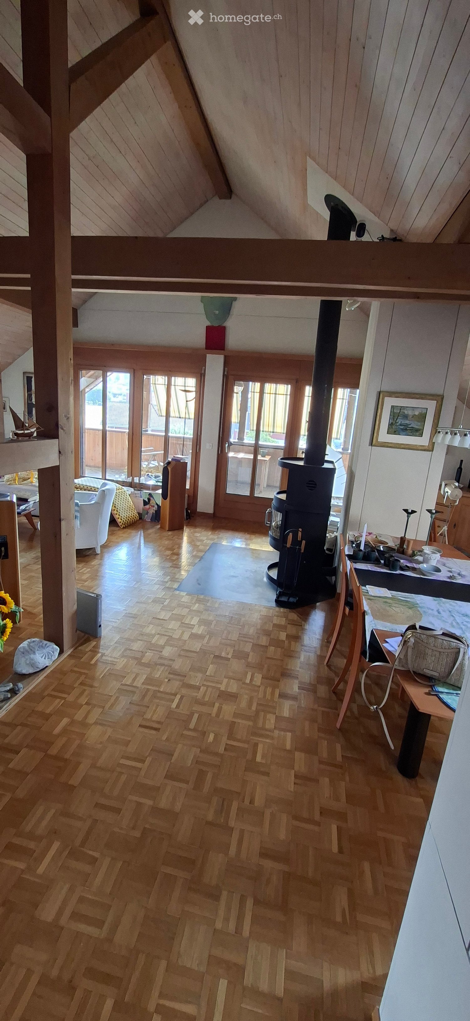 Bright, open living space with wooden herringbone flooring, white walls, and large windows. Features a wood stove, sofa, dining table with chairs, and various decorative items including a picture frame and a sunflower.