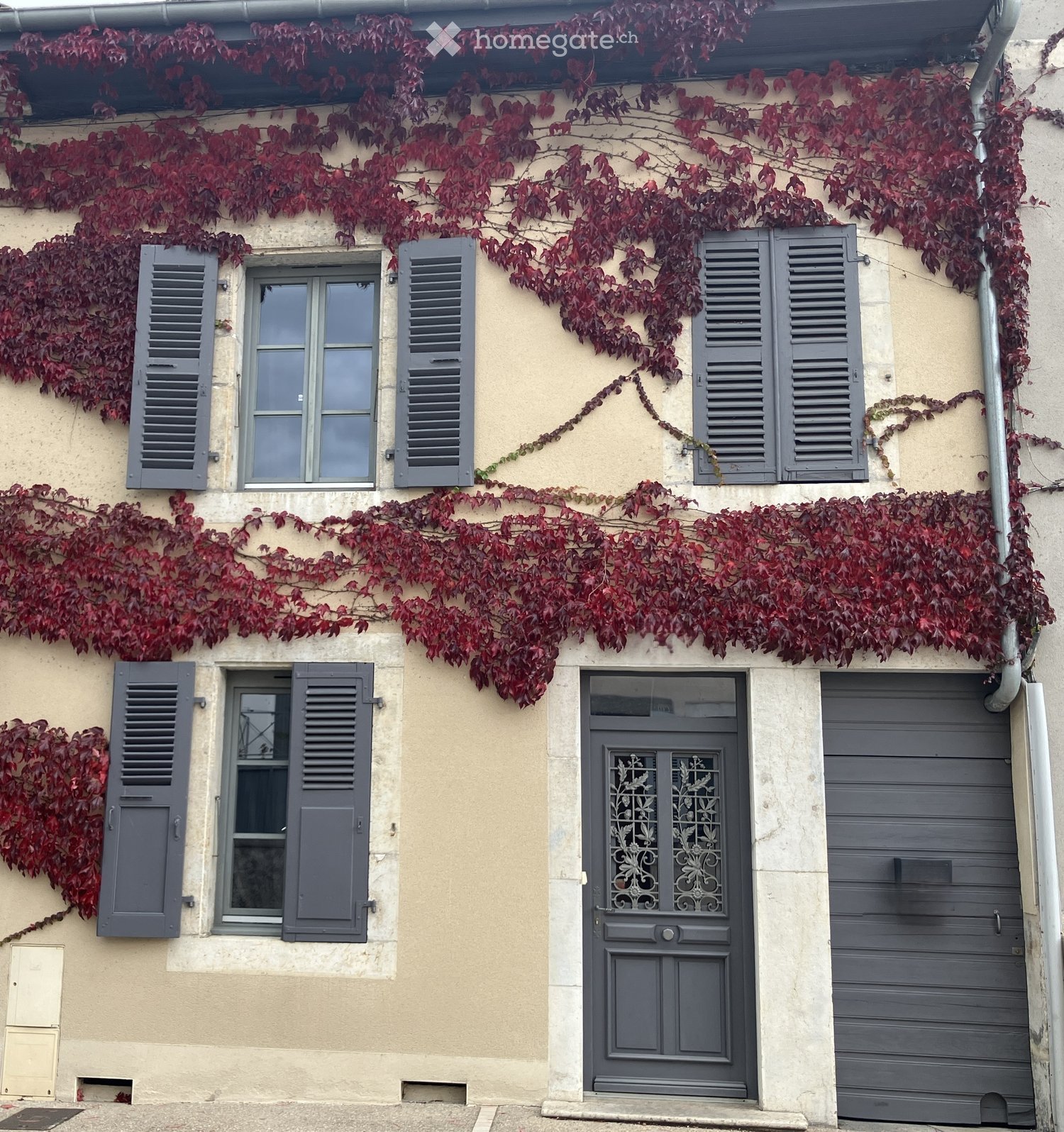 Two-story building with beige walls, grey shutters, and a grey garage door. Red climbing vines cover the facade, and a decorative grey front door with ornate glass panels is visible.