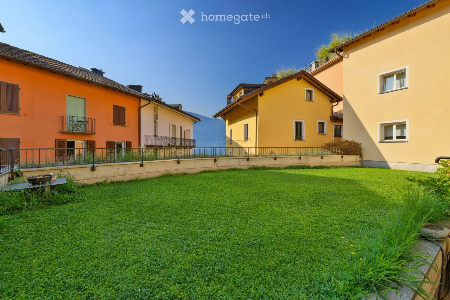 A well-maintained lawn in front of colorful residential buildings with balconies and railings, overlooking a scenic mountain view.