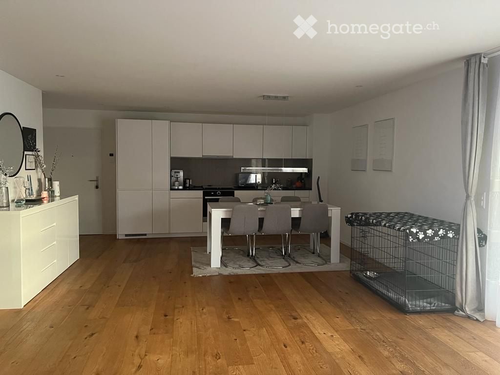 Modern kitchen and dining area with light-colored cabinets, a white dining table with four chairs, a mirror on the wall, and a pet cage with a blanket on top.