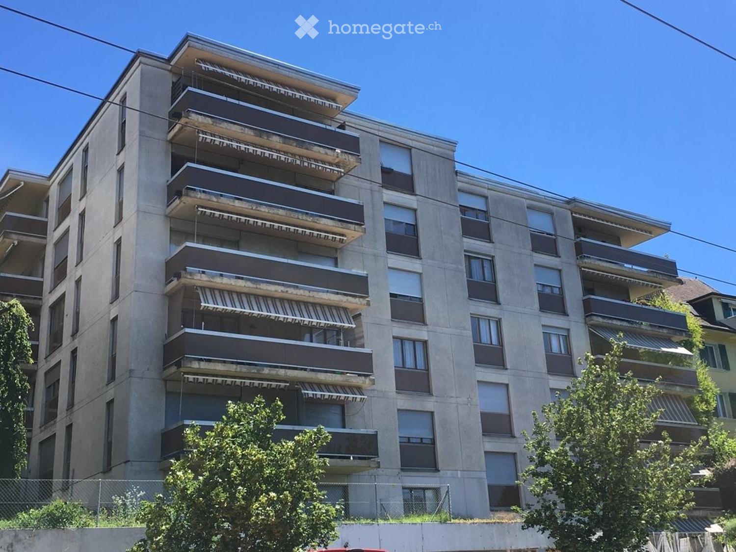 concrete apartment building, balconies, multiple floors, windows, plants, fenced area, trees