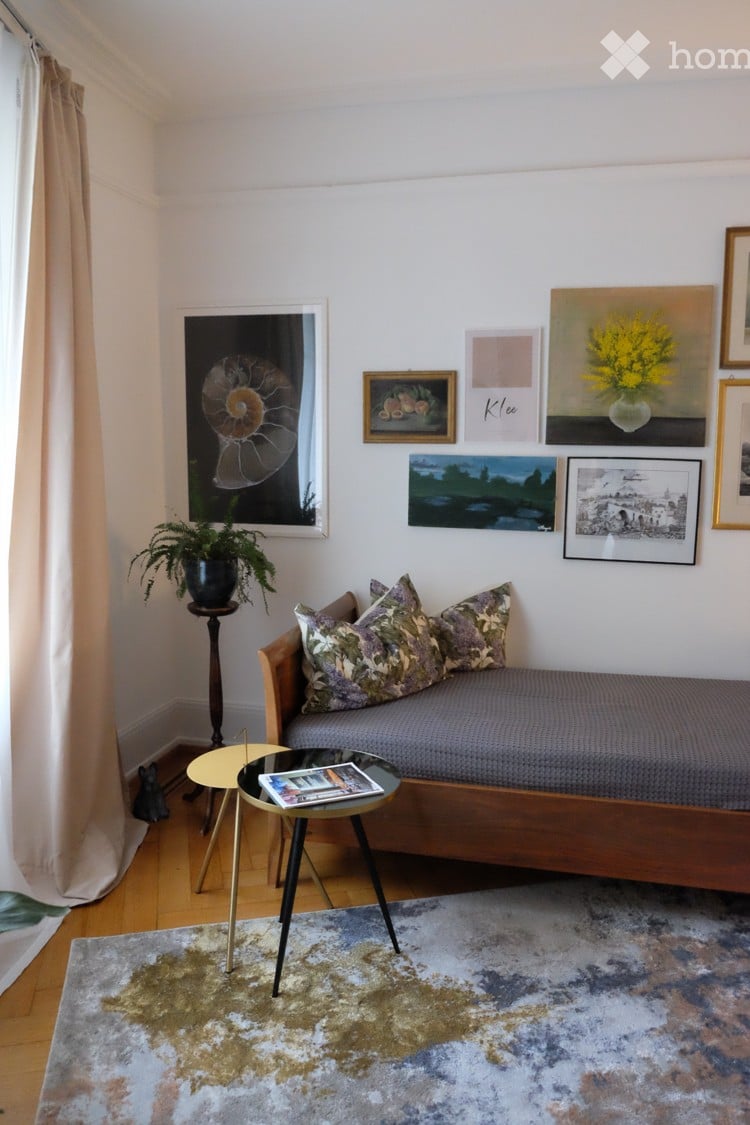 Cozy living room with a wooden sofa, patterned cushions, a small coffee table, a potted plant, and several picture frames on the wall.