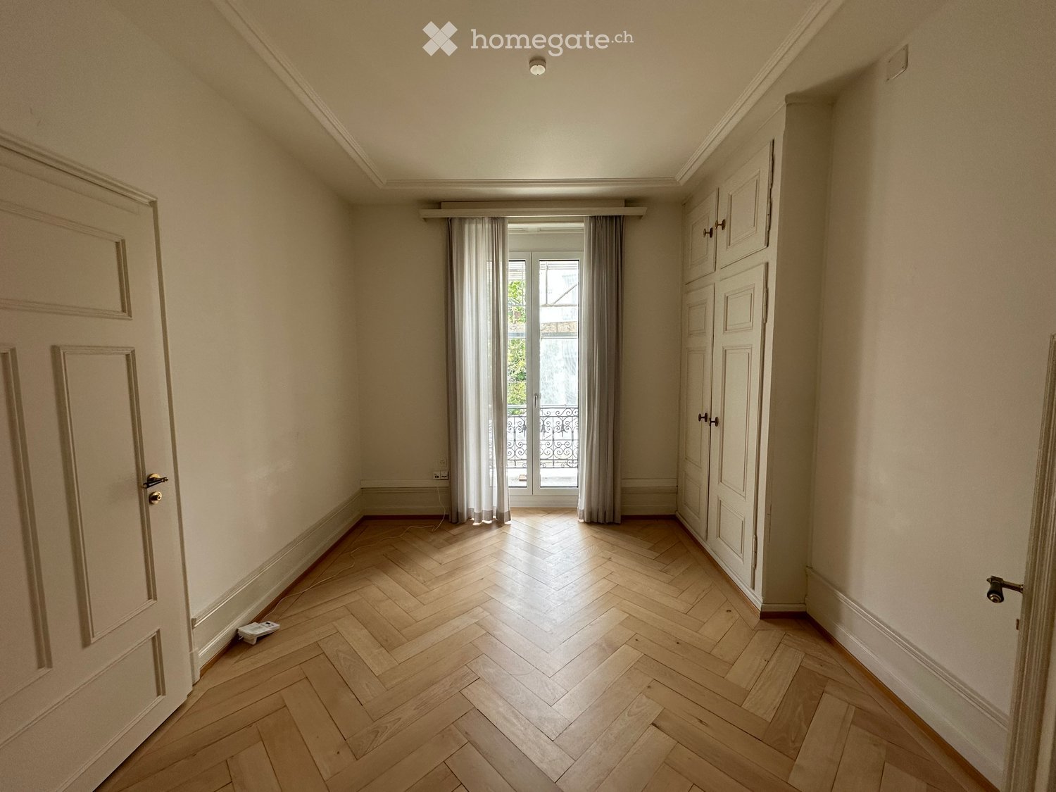 Empty room with wooden herringbone flooring, white walls, and a large window with curtains leading to a balcony.