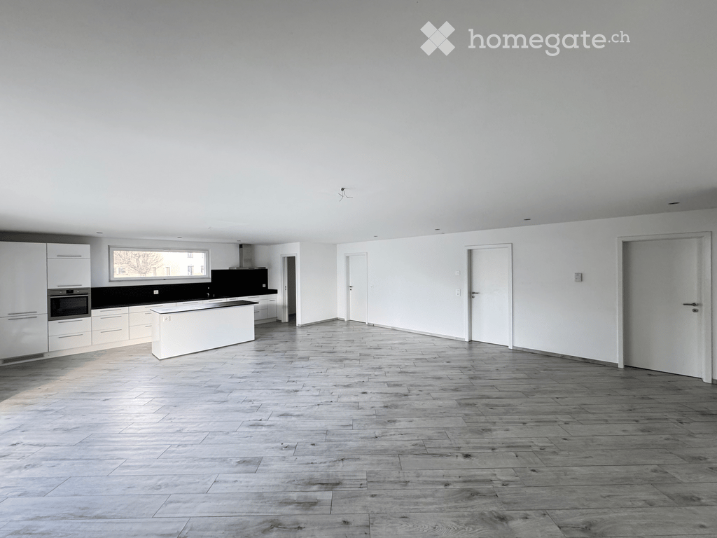 Open-plan space with a modern kitchen area featuring white cabinets, a central island, and a black countertop. The main area has a wooden floor and white walls, with several doors leading to other rooms.