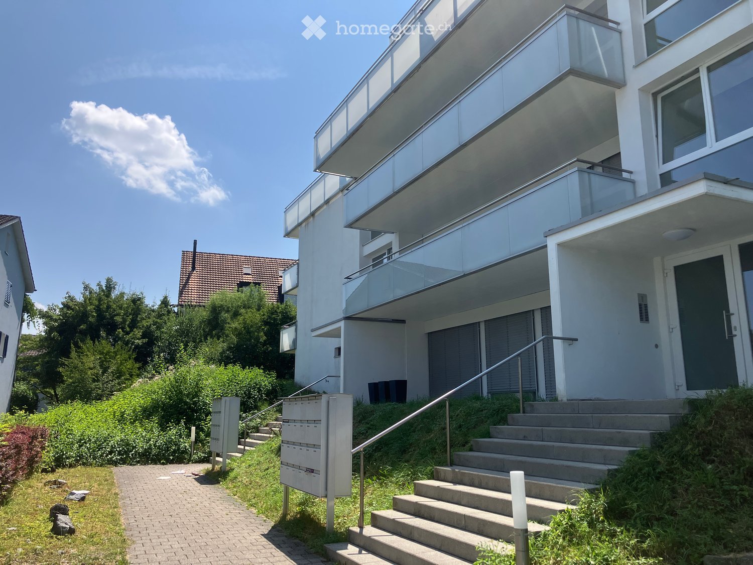 Modern two-story house with white facade, large windows, and glass balconies. Stairs lead up to the entrance.