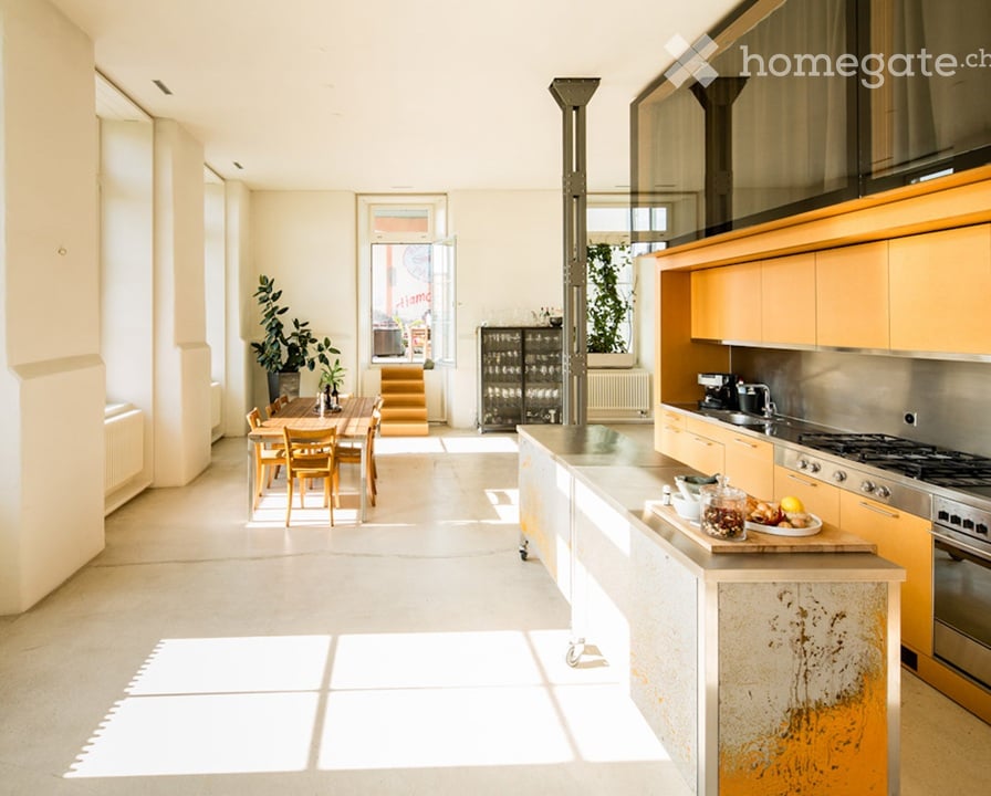 Bright, open kitchen with light-colored cabinets, stainless steel appliances, and a central island. A dining area with a wooden table and chairs is visible, along with potted plants and a window letting in natural light.