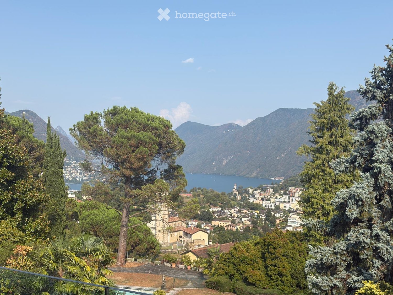 Panoramic view of a town with mountains and a lake in the background, surrounded by trees and greenery.