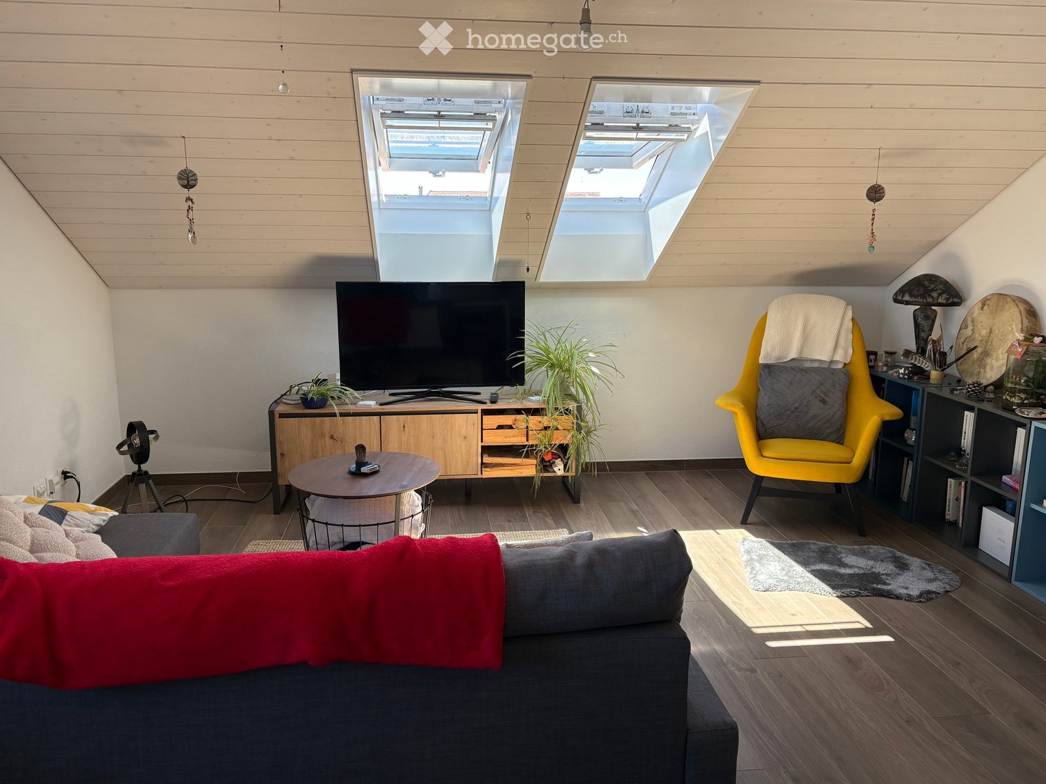 Modern living room with wooden floors, slanted ceiling, skylights, grey sofa, coffee table, yellow armchair, TV, and decorative plants.