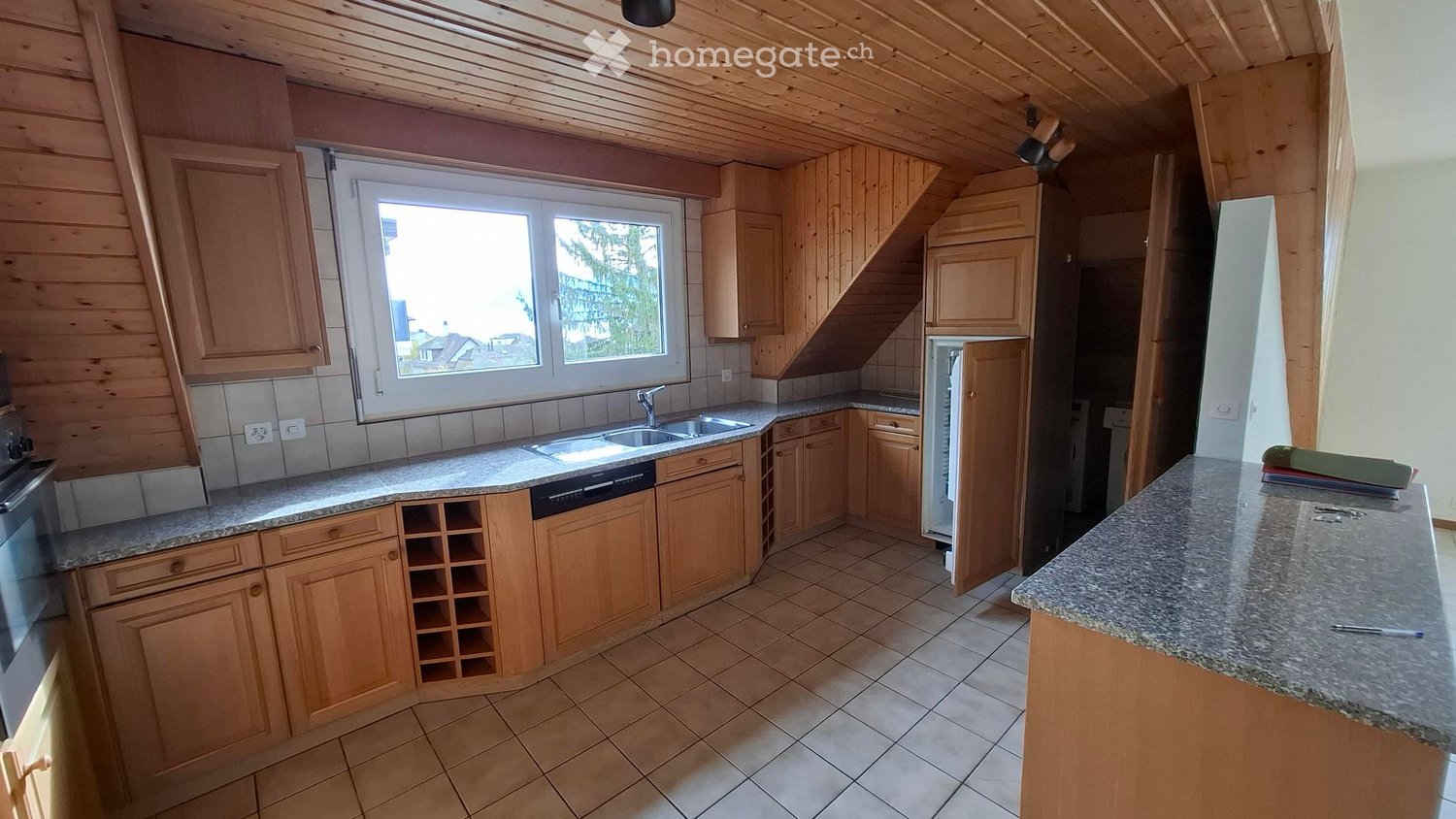 Bright kitchen with wooden cabinets, granite countertops, and a large window overlooking the outdoors. Includes a sink, oven, refrigerator, and a wine rack.