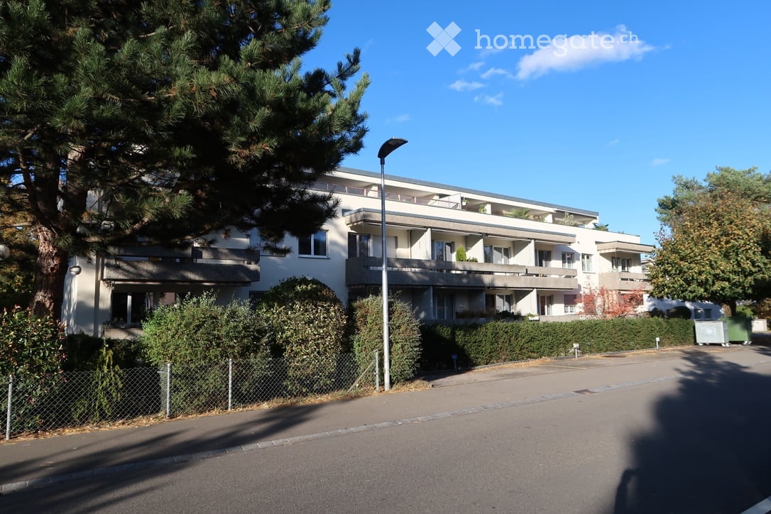 Modern multi-story apartment building with balconies, surrounded by greenery and a well-maintained hedge. The building features large windows and a clean, contemporary design.
