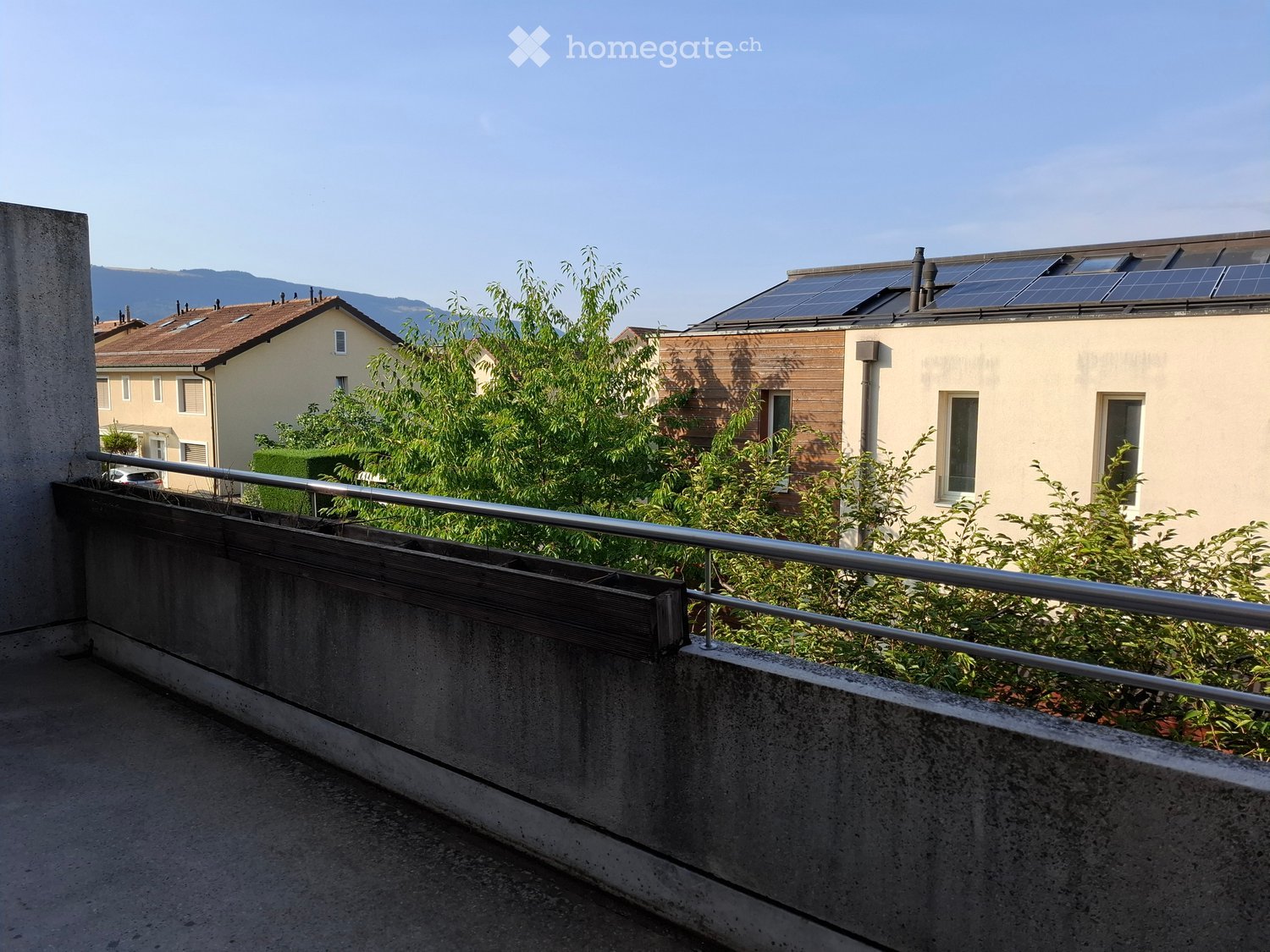 Balcony with metal railing, concrete edge, and greenery in the background, overlooking a residential area with houses and solar panels on the roof.