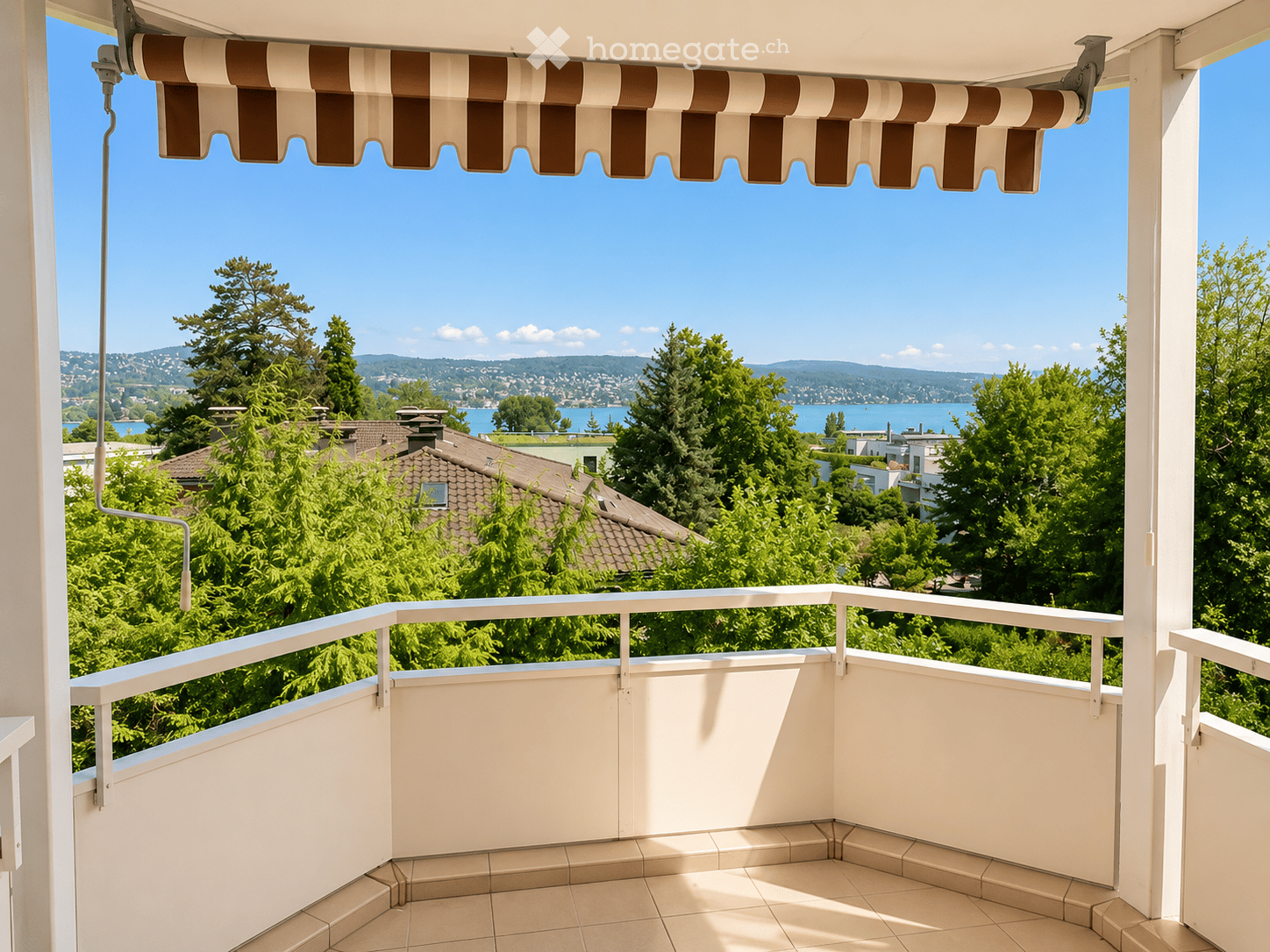 Balcony with white railings, tiled floor, and striped awning, overlooking a scenic lake and distant hills.
