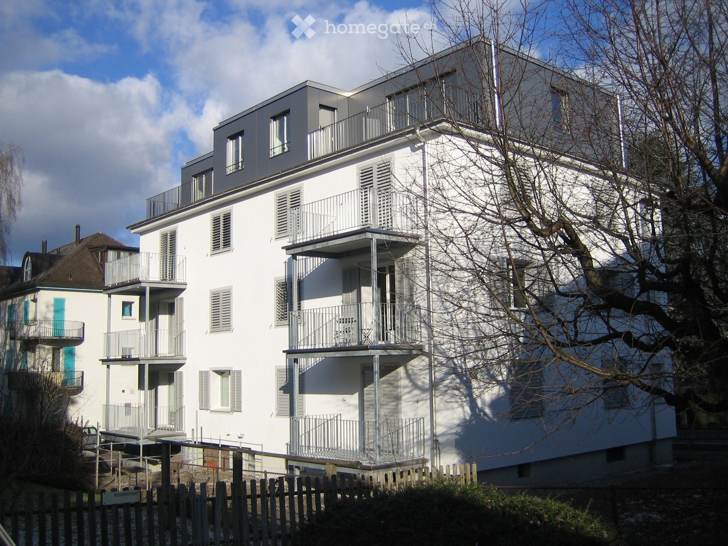 Modern white multi-story building with multiple balconies and large windows, surrounded by trees and a wooden fence.