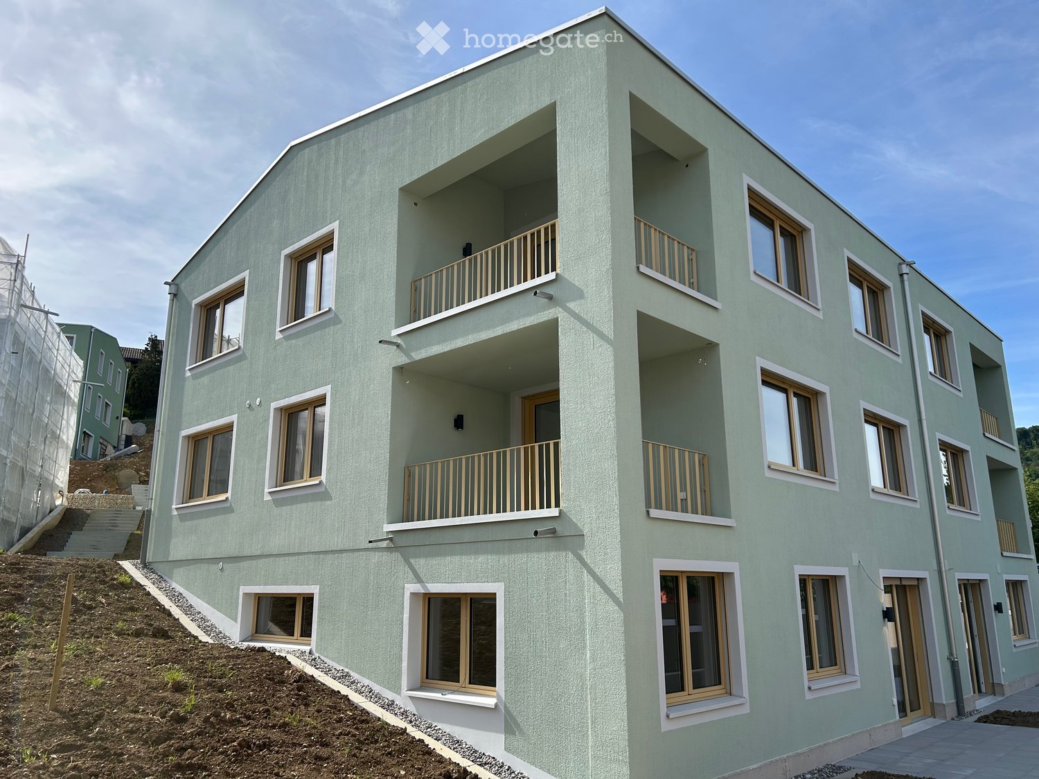 Modern multi-story building with light green facade, multiple windows, and balconies with wooden railings.