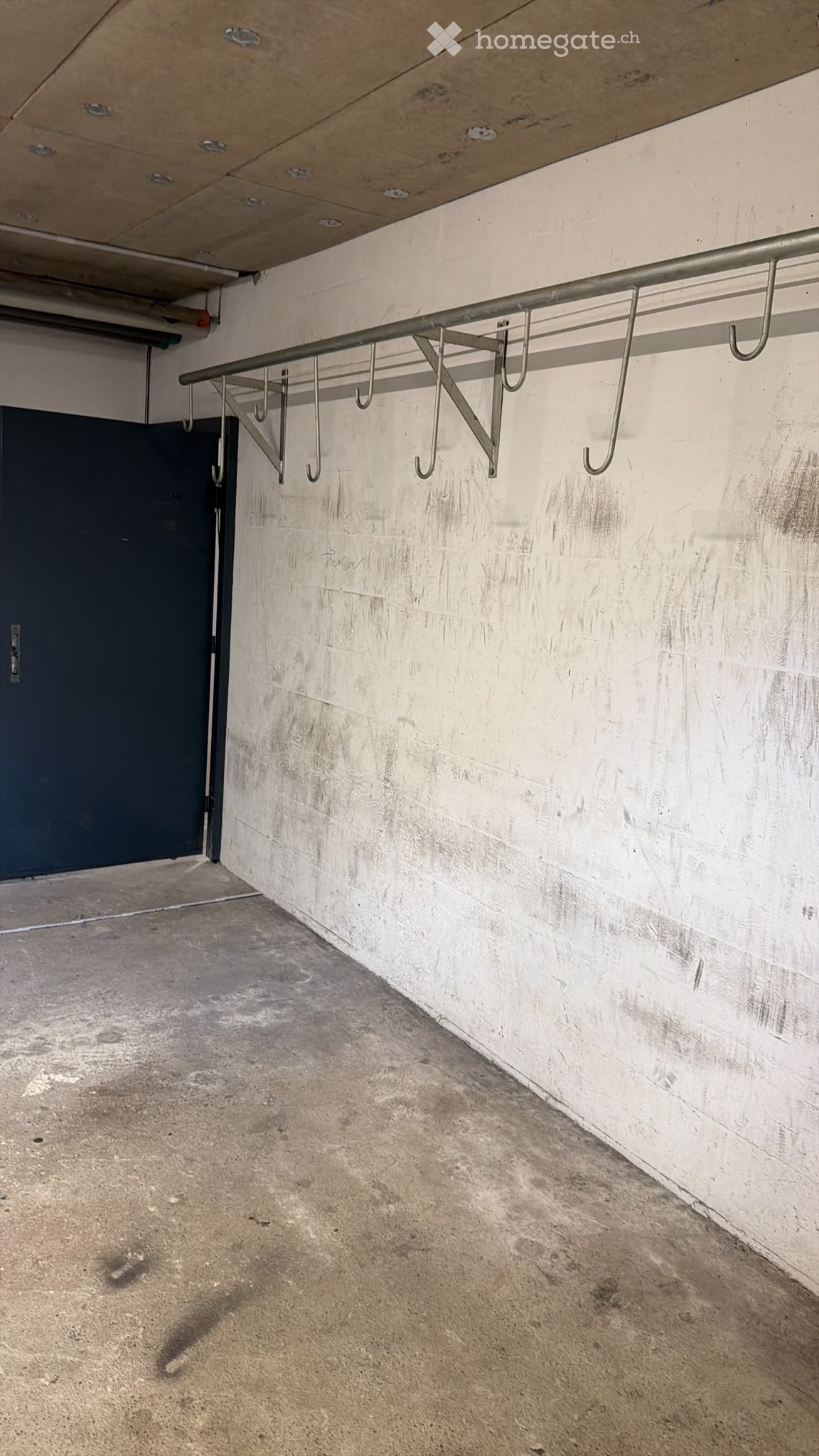 Empty concrete-floored space with a metal coat rack attached to a white brick wall and a dark blue door visible in the background.