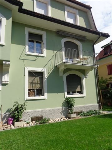 A two-story house with light green exterior walls, white-framed windows, and a balcony on the upper floor. The front yard features a well-maintained lawn, small garden beds with plants, and decorative rocks along the base of the house.