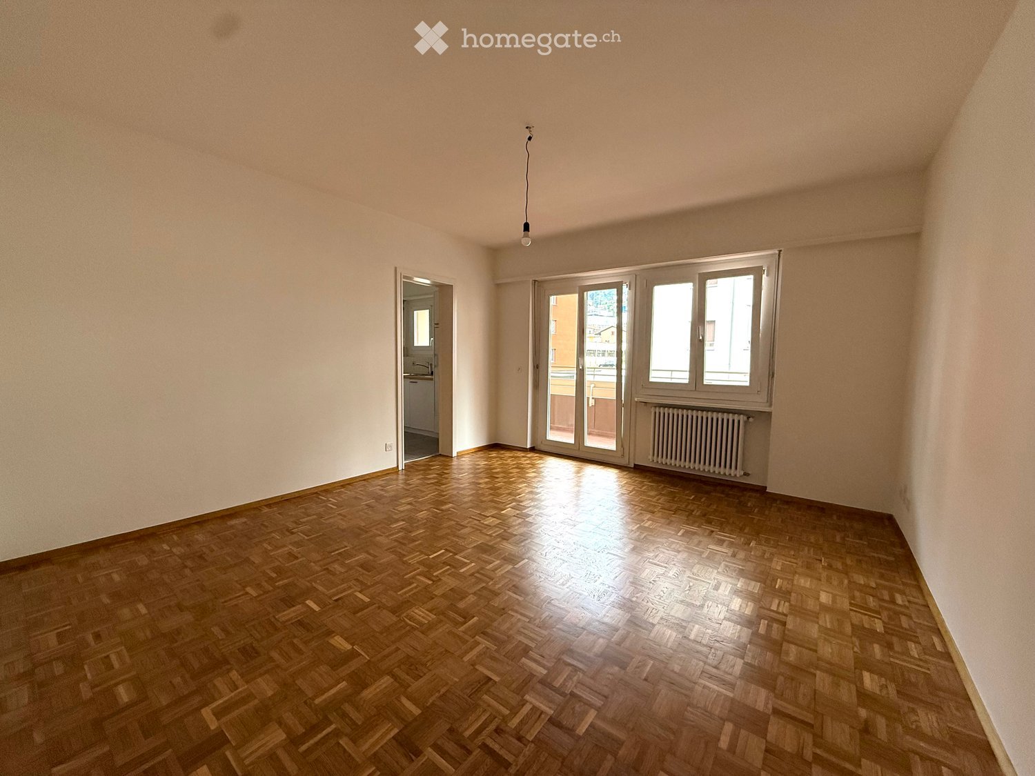 Empty room with wooden herringbone flooring, white walls, large windows, and a radiator. No furniture or equipment present.