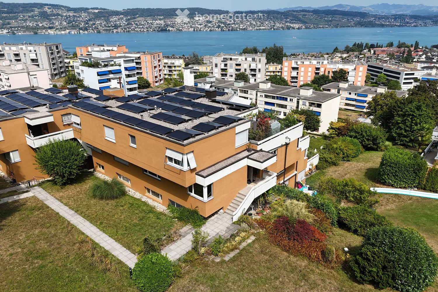 A modern, multi-story house with solar panels on the roof, surrounded by greenery and a well-maintained lawn. The house features large windows and a balcony, with a scenic view of a lake and distant mountains in the background.