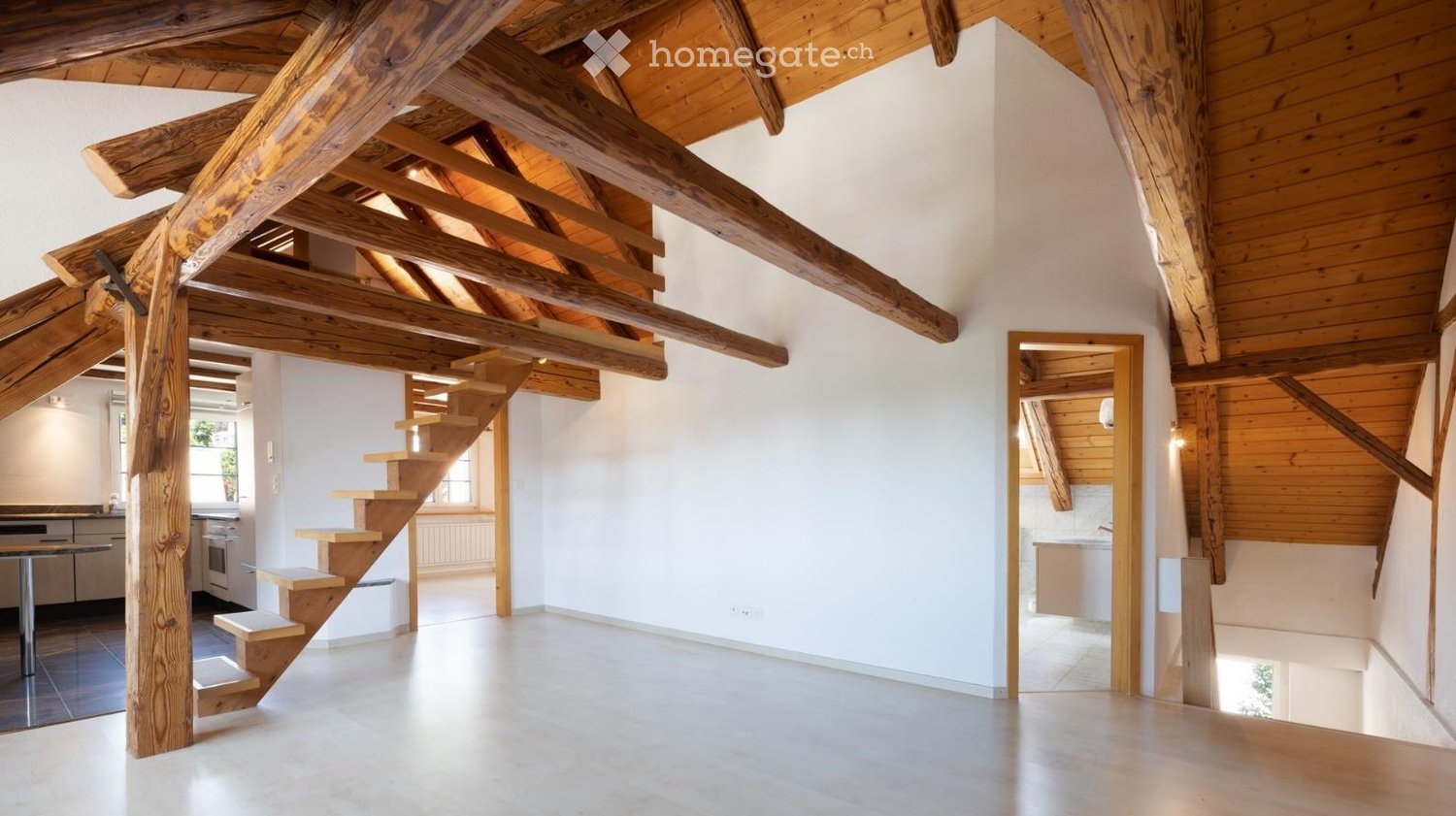 Bright, open loft space with exposed wooden beams and a wooden staircase leading to an upper level. The room features white walls, a smooth concrete floor, and large windows allowing natural light to fill the space.