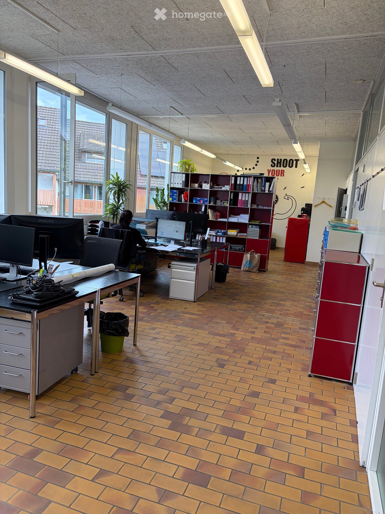 Office space with wooden tiled floor, desks, chairs, computers, shelves, and plants near large windows.