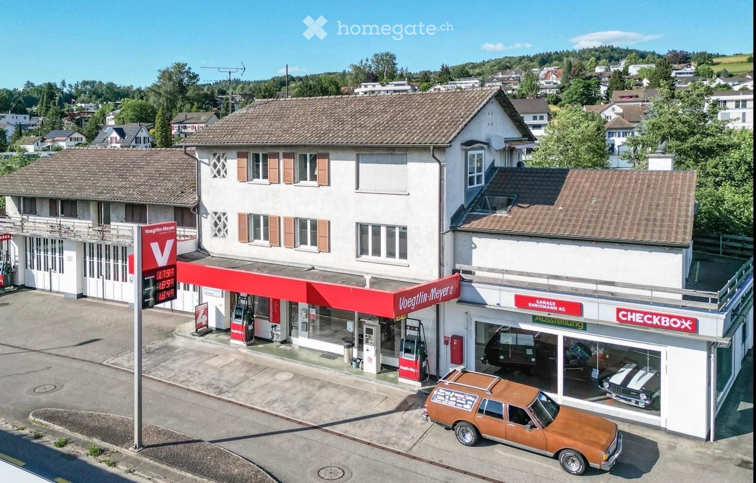 A commercial building with a gas station and car showroom, featuring a red canopy, gas pumps, and a display of vintage cars.