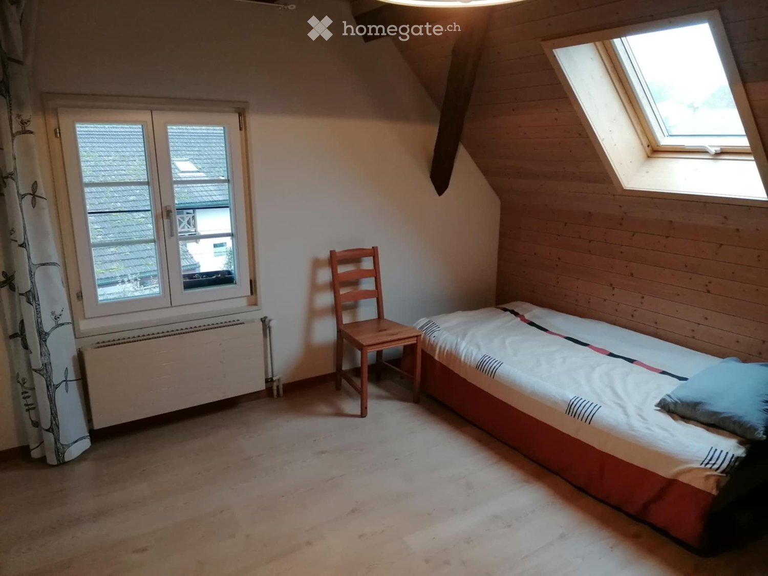 Cozy attic bedroom with wooden walls, slanted ceiling, skylight window, and a single bed with a patterned duvet and pillows. A wooden chair is placed next to the bed.