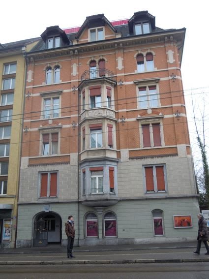 A multi-story building with a mix of orange brick and grey stone facade, featuring numerous windows with red shutters, ornate balconies, and decorative architectural details.