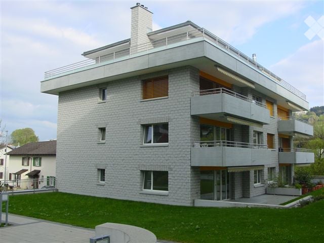 Modern multi-story building with grey stone facade, multiple balconies, large windows, and a chimney on the roof.