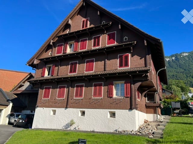 Traditional Swiss-style house with brown wooden facade, red shutters, and white lower section. Two-story structure with multiple windows and a pitched roof.