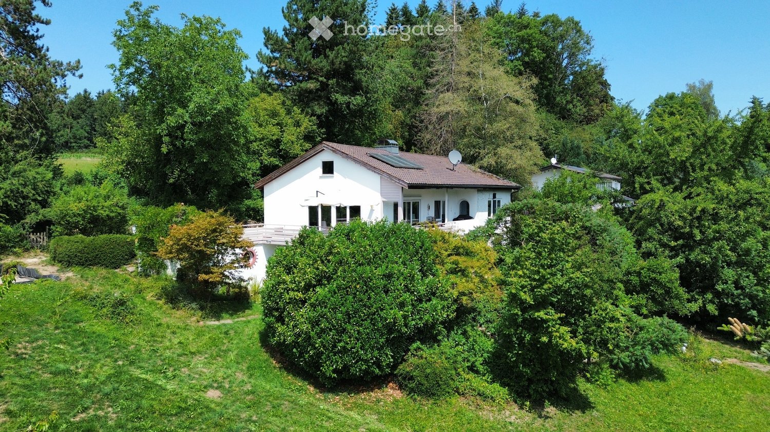 A two-story white house with a red-tiled roof, surrounded by lush greenery and trees. The house features large windows, a balcony, and a solar panel on the roof.