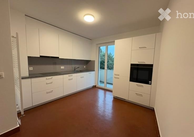 Modern, minimalist kitchen with white cabinets, a dark countertop, and a built-in oven. Features a large window with a sliding glass door leading to a balcony.