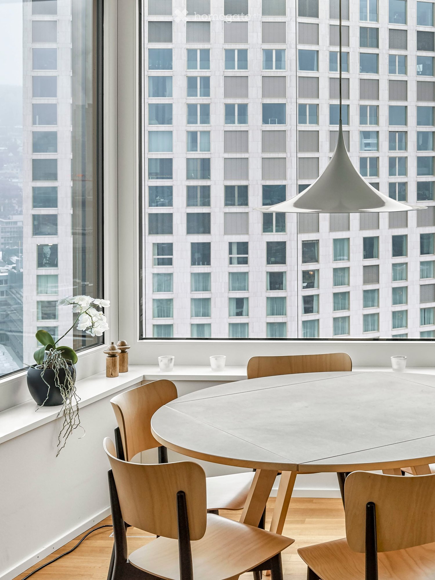 Dining area with a round table, wooden chairs, a white wall, a window with a view of the city, a white lamp, and a potted plant on the window sill