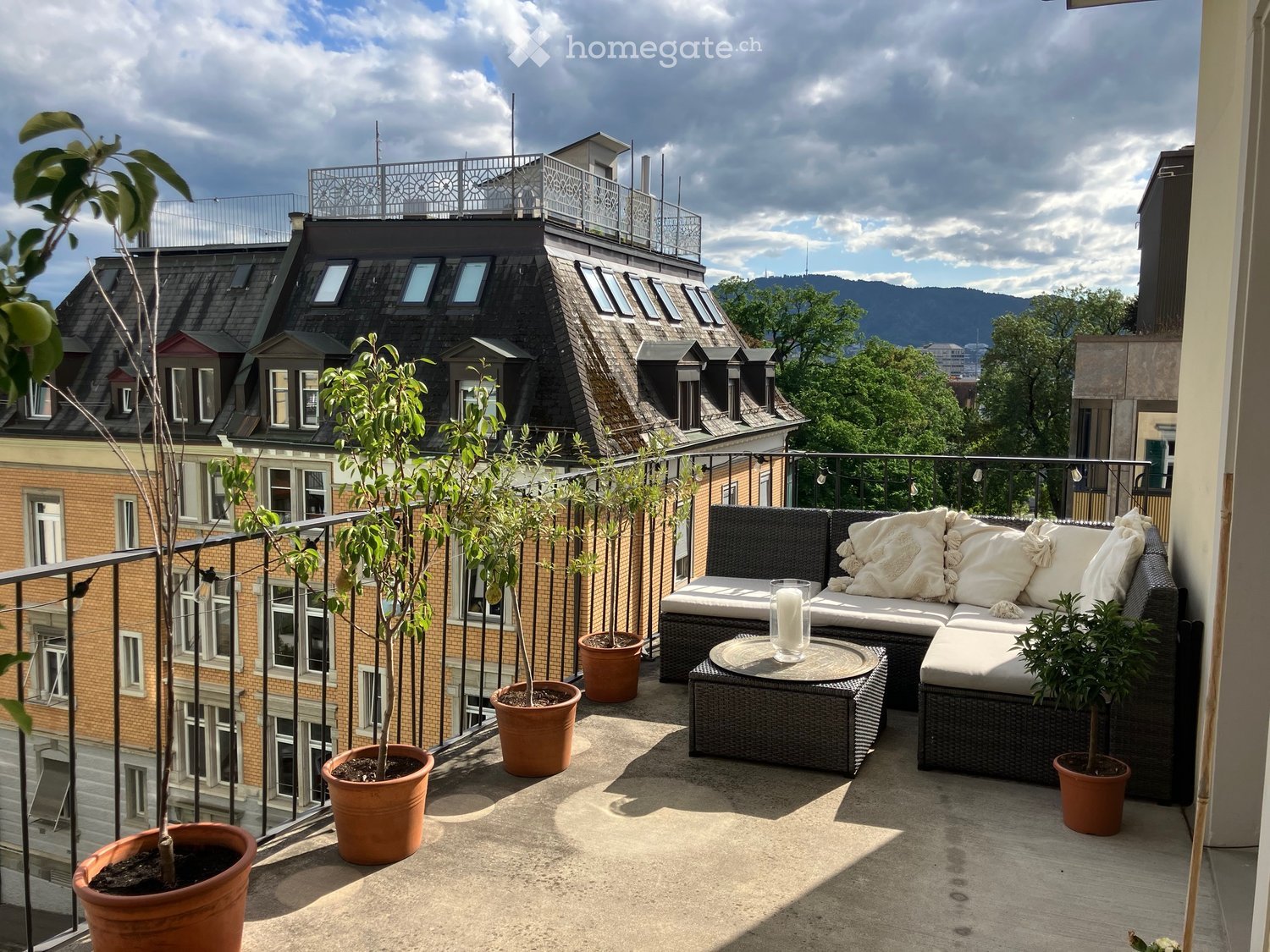 Balcony with a modern wicker sofa, coffee table, and several potted plants, overlooking a cityscape with rooftops and mountains in the background.